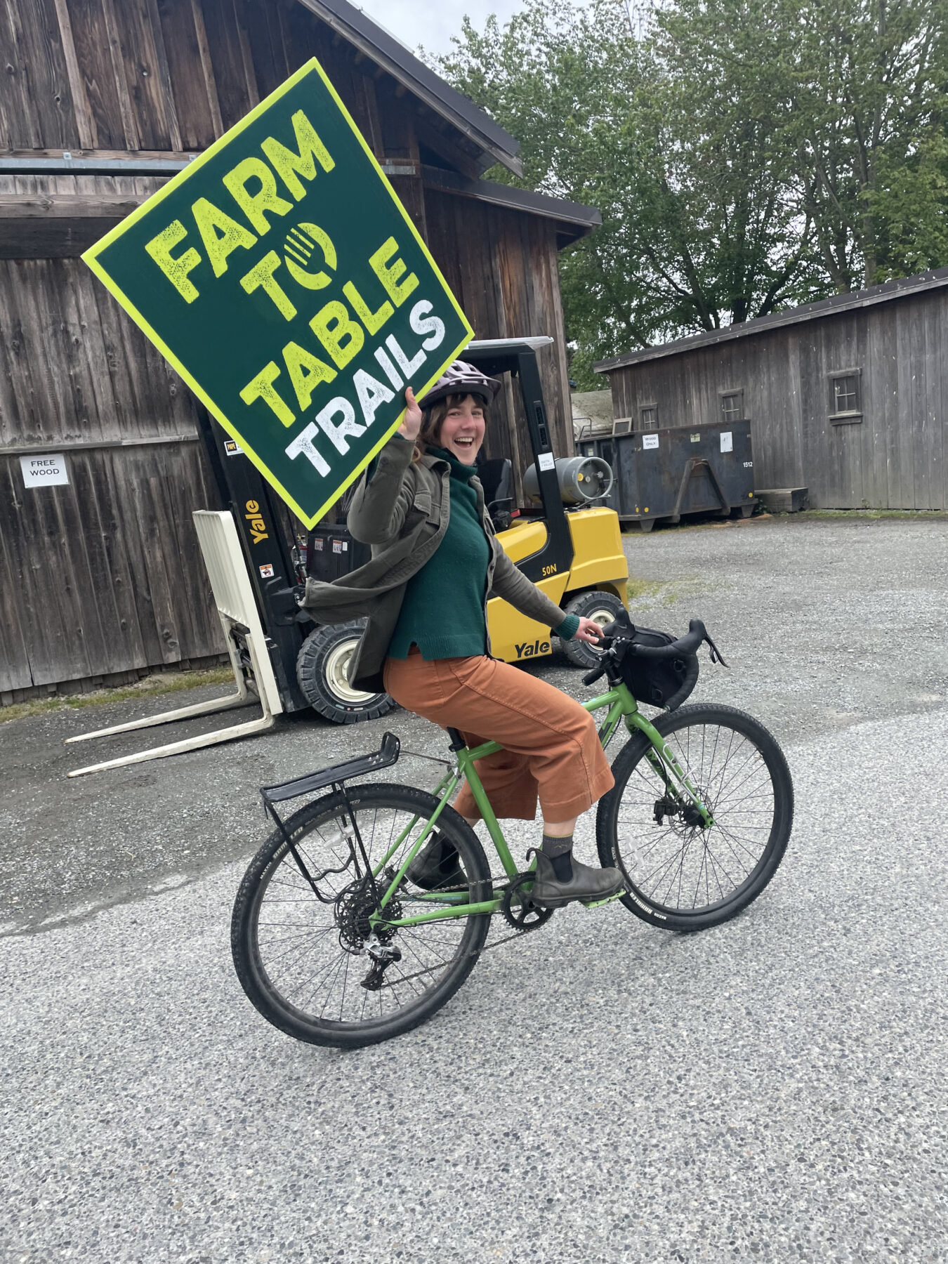 20250516_200745178_iOS (1) a woman riding a bike holds up a sign that says farm to table trails