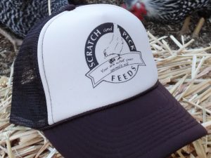 A black and white trucker hat with the Scratch and Peck Feeds logo on a bed of hay with hens in the background.